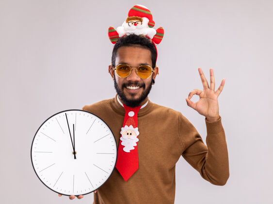 高兴非洲裔美国人 身穿棕色毛衣 头戴圣诞老人耳环 打着有趣的红色领带 拿着时钟看着摄像机 兴高采烈地微笑着 白色背景上立着ok标志头搞笑领带