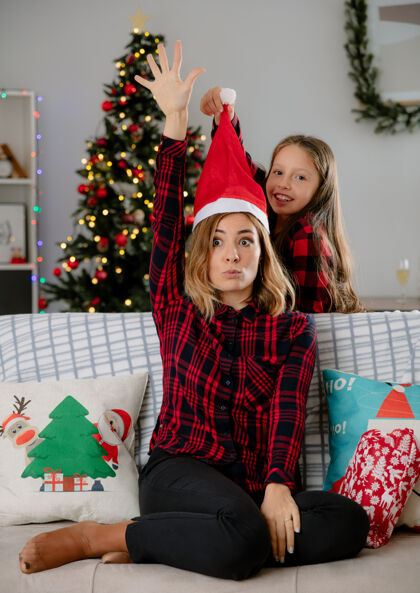 头微笑的女儿把圣诞帽戴在妈妈头上 举起手坐在沙发上 在家里享受圣诞时光坐抚养女儿