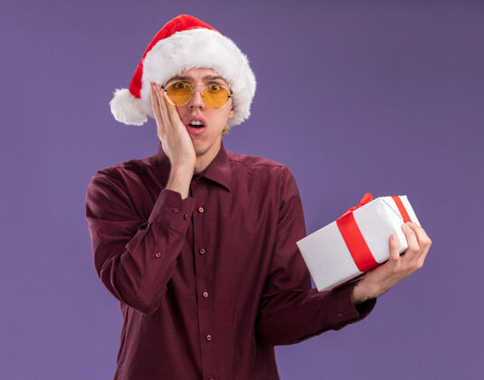 保持戴着圣诞帽戴着眼镜拿着礼包看着相机手放在紫色背景上孤立的脸上的年轻金发男人帽子眼镜关心
