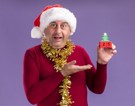 二十中年男子戴着圣诞老人帽子 脖子上戴着金属丝 手里拿着日期为25的玩具立方体 手举着微笑站在紫色背景上中间五举行