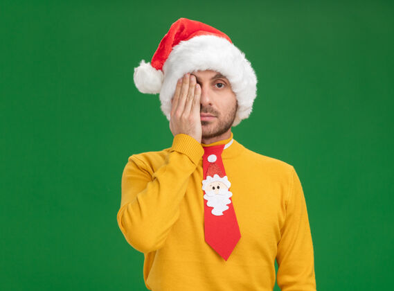 年轻年轻的白人男子戴着圣诞帽 打着领带 用手遮住半张脸 看着隔离在绿色背景上的相机 还有复印空间一半相机圣诞节