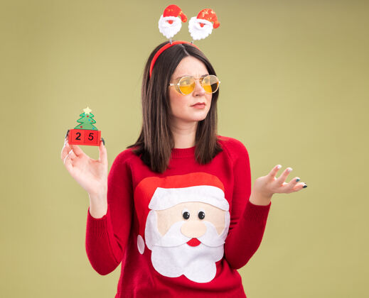 皱眉皱眉年轻漂亮的白人女孩穿着圣诞老人毛衣和戴眼镜的头带拿着圣诞树玩具和日期看一边显示空手孤立在橄榄绿的背景女孩圣诞老人圣诞老人