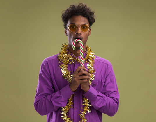 嘴令人印象深刻的非洲裔美国年轻男子戴着眼镜 脖子上戴着金箔花环 嘴里拿着圣诞糖果手杖 看着橄榄绿背景上孤立的摄像机眼镜举行手杖