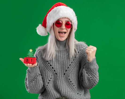圣诞老人年轻的金发女郎穿着冬季毛衣 戴着圣诞老人帽 戴着红色眼镜 手里拿着玩具立方体 手里握着新年快乐的日期 紧握着拳头 站在绿色的背景上 快乐而兴奋金发眼镜站着