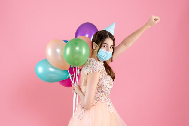面具戴着派对帽和医用面具的快乐女孩站成超级英雄的姿势 背着粉色的彩色气球背抱着粉色