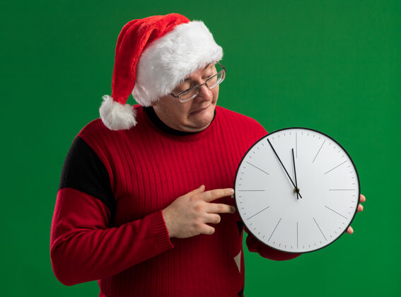 着急焦虑的成年男子戴着眼镜 戴着圣诞老人的帽子 拿着并看着绿色背景上孤立的时钟大人帽子钟
