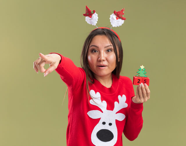 亚洲人惊讶的年轻亚洲女孩戴着圣诞发箍拿着圣诞玩具点在相机上孤立的橄榄绿背景箍点举行