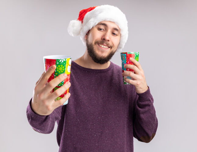 微笑一个穿着紫色毛衣 戴着圣诞帽的年轻人 手里拿着五颜六色的纸杯 站在白色的背景上兴高采烈地看着纸杯圣诞老人高兴杯子