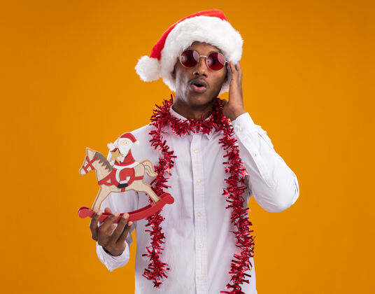 眼镜一个年轻的非洲裔美国人 戴着圣诞帽 戴着眼镜 脖子上戴着金箔花环 抱着摇摇木马上的圣诞老人 看着摄像机 抚摸着孤立在橙色背景下的头金属片举行周围