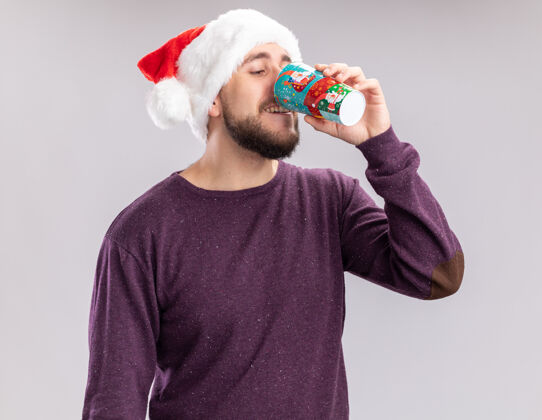 年轻人穿着紫色毛衣 戴着圣诞帽 站在白色背景上 用彩色纸杯喝酒的快乐年轻人喝酒圣诞老人杯子