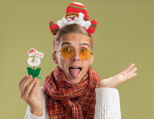 手可爱的年轻帅哥戴着圣诞老人的头带和围巾看着相机拿着雪人玩具显示空手和舌头孤立在橄榄绿的背景舌头年轻橄榄