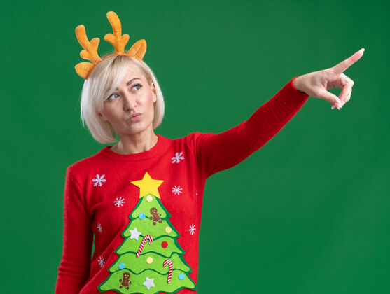自信自信的中年金发女人 戴着圣诞驯鹿鹿角头带和圣诞毛衣 一边看着一边指着绿色背景上孤立的噘嘴噘嘴中年指尖