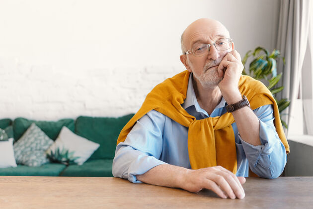 家一张室内照片 60岁的退休老人 留着白胡子 光头 坐在客厅的木桌旁 无聊地 手放在下巴上 神情若有所思老人看客厅