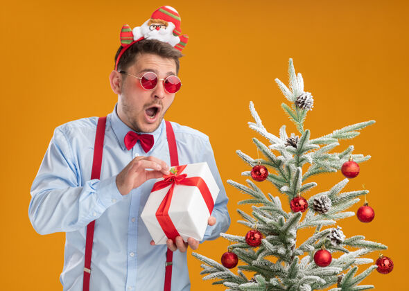 站立戴着吊带领结的年轻人站在圣诞树旁 戴着圣诞老人和红眼镜 手里拿着一份礼物 在橙色的背景下快乐而兴奋轮辋圣诞老人举行