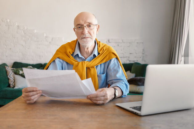 休闲人 财务 技术和工作观念严肃时尚的退休商人在现代办公室做财务工作 手里拿着文件 打开木桌上的笔记本电脑从他身边财务桌子办公室