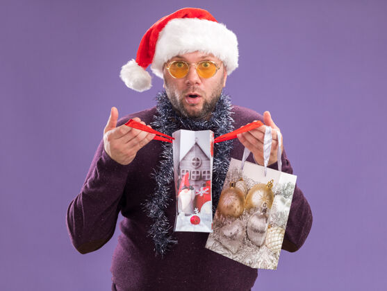 礼物好奇的中年男子戴着圣诞帽 脖子上戴着金属丝花环 戴着眼镜 手里拿着圣诞礼品袋 打开一个 看着紫色背景上孤立的相机包好奇举行