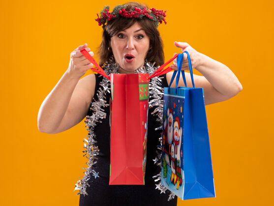 头好奇的中年妇女头戴圣诞花环 脖子上戴着金属丝花环 手里拿着圣诞礼品袋 打开一个 看着孤立在橙色背景上的相机包周围金属丝
