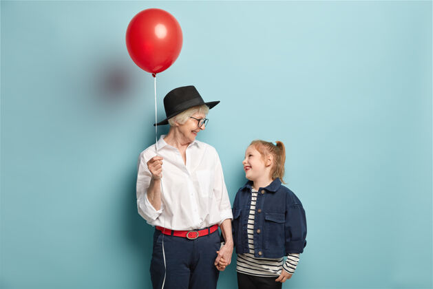 团结摄影棚拍摄的快乐小孩用气球牵着奶奶的手 互相看着 度过自由时光眼镜帽子一代
