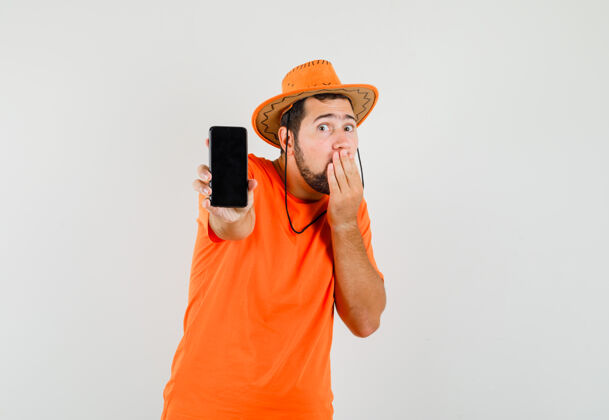 男性年轻人展示手机在橙色的t恤 帽子和期待惊讶 前视图手势休闲表情