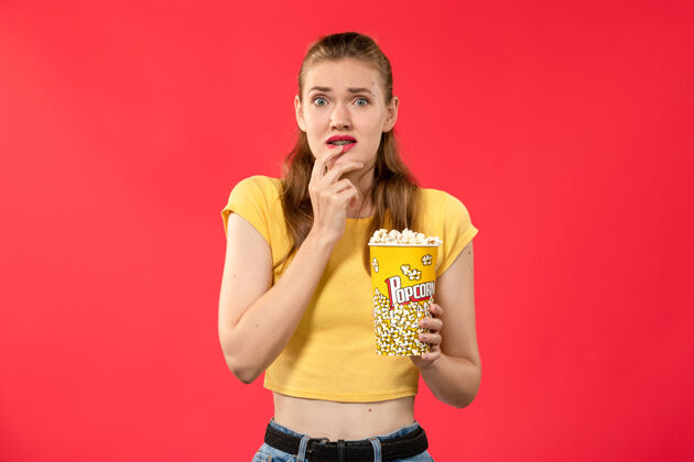 电影院前视图年轻女性拿着爆米花包 带着恐惧的表情出现在淡红色的墙壁电影院里成年人包装音乐家