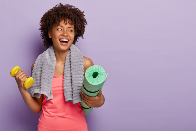 健身积极的黑皮肤女人拿着瑜伽垫和哑铃 穿着运动服 脖子上有毛巾举重运动发型