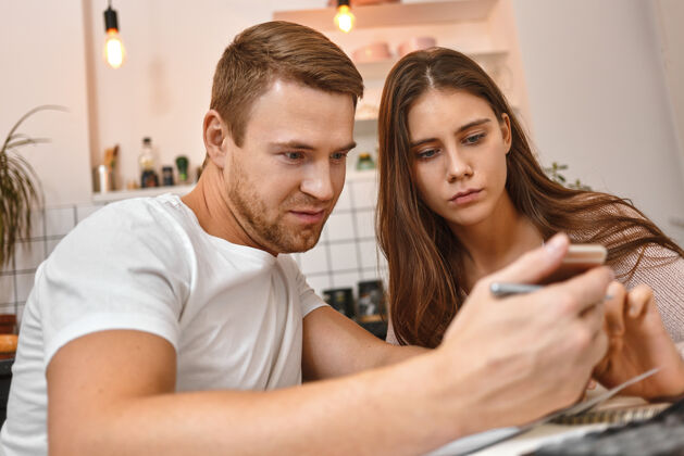 科技一对年轻夫妇在厨房喝咖啡 用手机阅读网上银行关于未付抵押贷款的通知 带着担忧的表情看着屏幕在一起家庭自拍