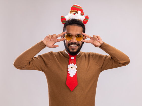 非洲一位非洲裔美国人 身穿棕色毛衣 头戴圣诞老人戒指 打着有趣的红色领带 面带微笑地看着摄像机 在白色背景下 眼睛附近有一个v型标志Vsign轮辋圣诞老人