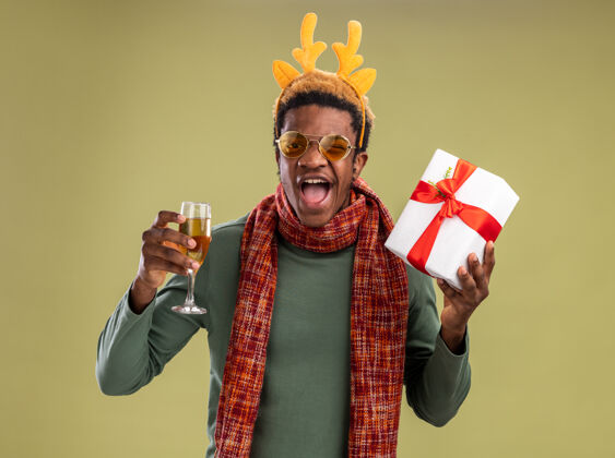 圣诞节一个非洲裔美国人 戴着滑稽的边 戴着鹿角 脖子上围着围巾 手里拿着一杯香槟和圣诞礼物 站在绿色的背景下 看着摄像机 他既高兴又兴奋非洲围巾礼物