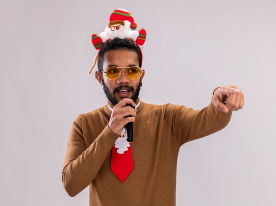 麦克风非洲裔美国人 身穿棕色毛衣 头戴圣诞老人戒指 打着有趣的红色领带 拿着麦克风 食指指向站在白色背景上的一边美国人圣诞老人边缘