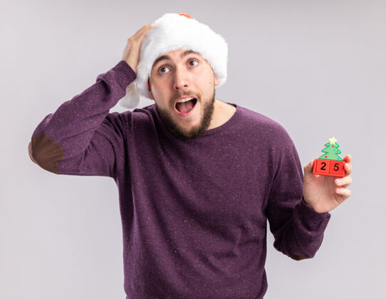 立方体穿着紫色毛衣 戴着圣诞帽的年轻人站在白色的背景上 一边看一边惊奇和惊讶地展示着方块和新年约会帽子惊讶惊讶