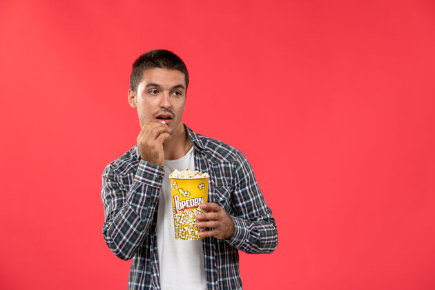 爆米花正面图年轻男子拿着爆米花包在浅红的墙上吃电影年轻人电影院音乐家