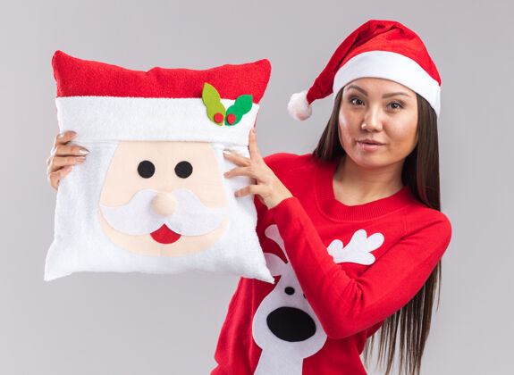 高兴高兴的年轻亚洲女孩戴着圣诞帽和毛衣拿着圣诞枕头隔离在白色背景上亚洲人年轻帽子