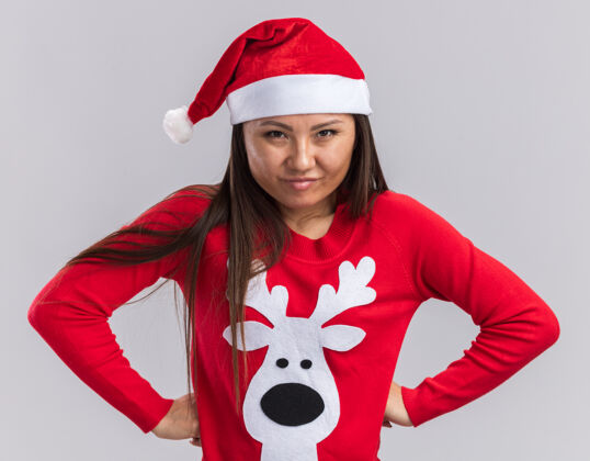 女孩自信的年轻亚洲女孩戴着圣诞帽 穿着毛衣 双手放在臀部 白色背景上 与世隔绝毛衣自信臀部