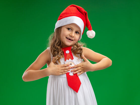 高兴高兴的歪着头戴着圣诞帽打着领带的小女孩把手放在自己的绿色背景隔离放女孩小