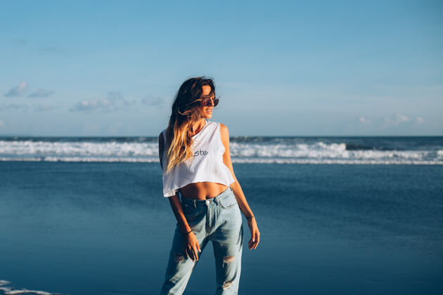 海洋美丽的白种女人 穿着白色上衣和牛仔裤 在日落的阳光下映照着海边健康裸体海滩