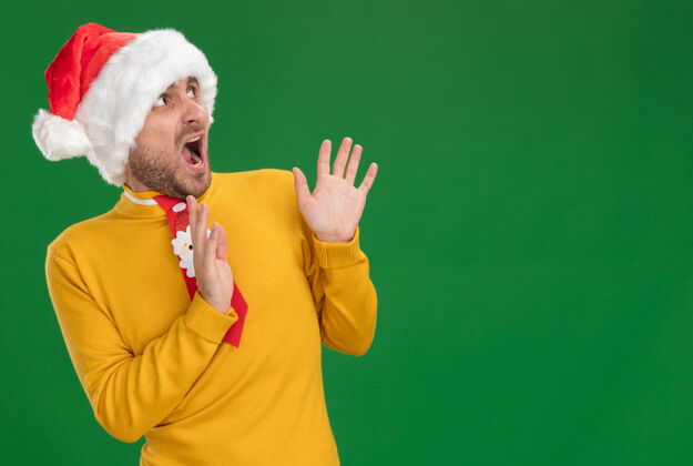 看一个戴着圣诞帽打着领带的年轻白种人惊恐地站在一边 双手举在空中尖叫着 在绿色的背景上隔离着 留着复制空间手空气帽子