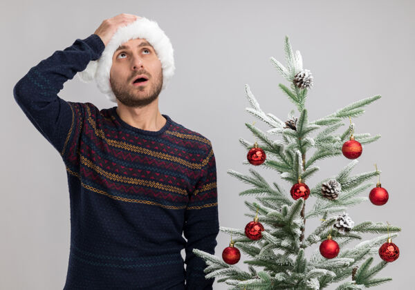 年轻遗憾的是 一个戴着圣诞帽的白人年轻人站在圣诞树旁抬头仰望 手放在头上 在白色背景下与世隔绝手头保持