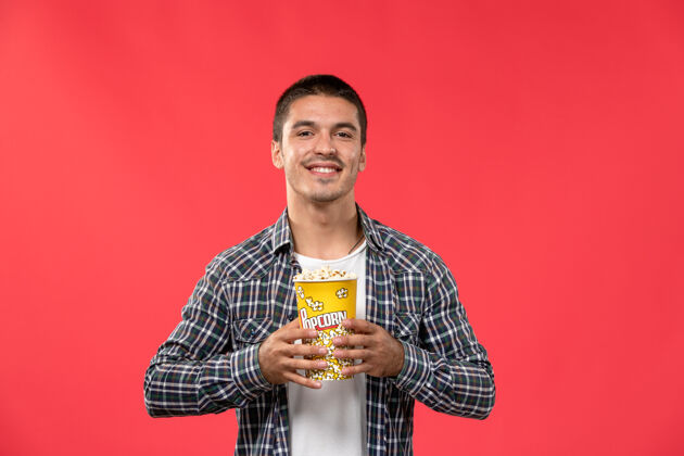 电影正面图年轻男子拿着爆米花包微笑在红墙电影院电影院包装年轻男性