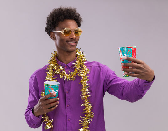 杯子微笑着的年轻的非裔美国人戴着眼镜 脖子上戴着金属丝花环 手里拿着塑料圣诞杯 伸出一个 孤立地看着它 背景是白色的伸展眼镜周围