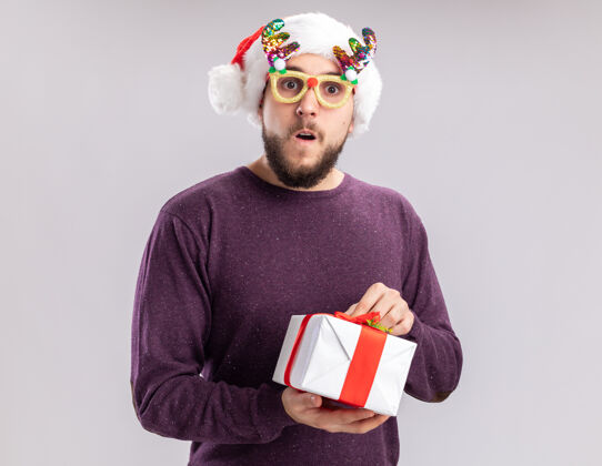 圣诞老人快乐的年轻人穿着紫色毛衣 戴着圣诞帽 戴着滑稽的眼镜 拿着礼物 站在白色的背景下惊讶地看着相机礼物惊讶毛衣