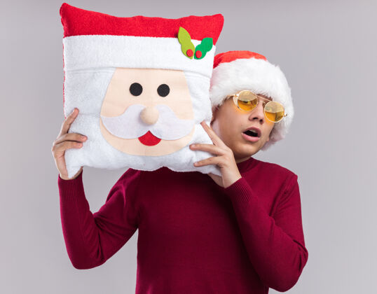 害怕害怕的年轻人戴着圣诞帽戴着眼镜拿着圣诞枕头隔离在白色背景上圣诞节抱着眼镜