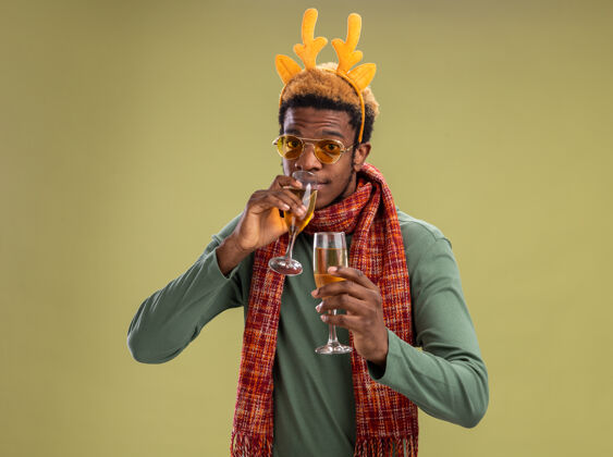 眼镜一个非洲裔美国人 他戴着鹿角 脖子上围着围巾 站在绿色的背景上喝着两杯香槟角站着拿着