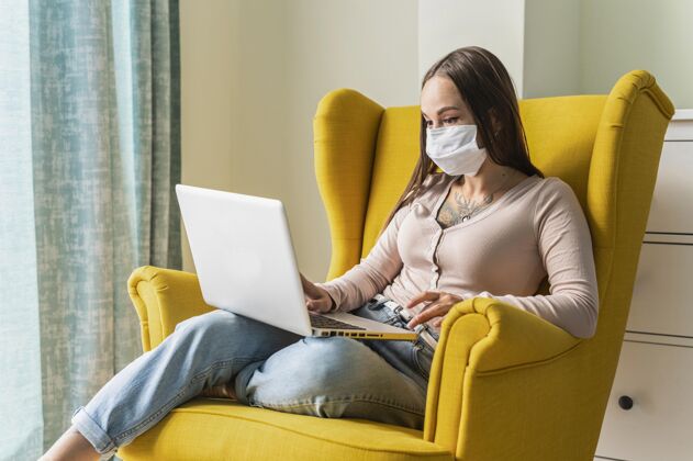 妇女大流行期间 戴着医用口罩 坐在扶手椅上操作笔记本电脑的妇女检疫社交口罩
