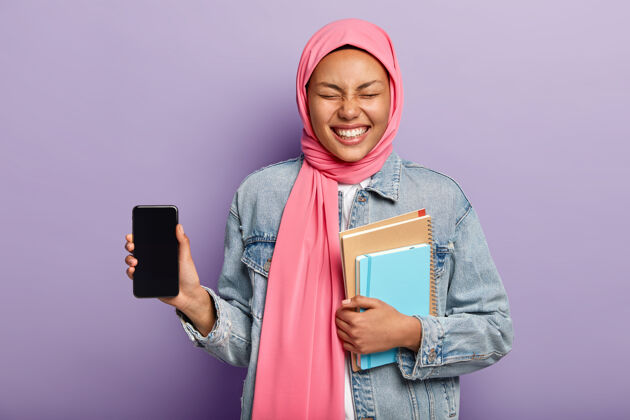 设备这是你需要的手机有伊斯兰观点的快乐女人 戴着传统的头巾 展示智能手机屏幕 大笑手机日记大笑