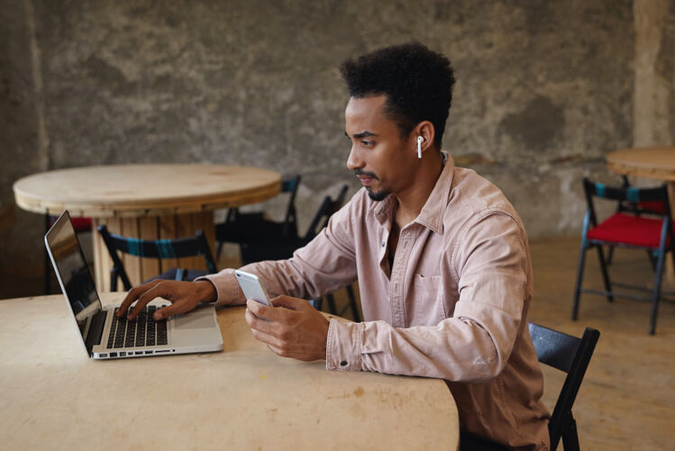 电话年轻漂亮的黑皮肤商人 留着胡子 在办公室外工作 坐在桌旁拿着笔记本电脑 穿着米色衬衫 拿着智能手机打电话男性20多岁衬衫