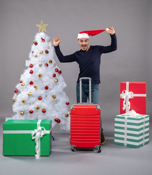 礼物站在白色圣诞树旁 手里拿着圣诞帽的年轻人欣喜若狂 礼物是灰色的拿着帽子礼物