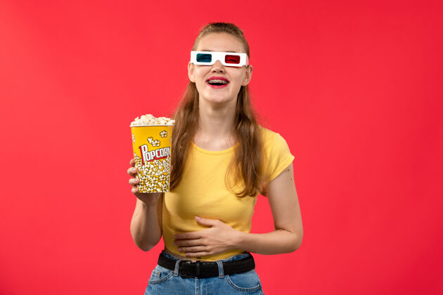 护目镜前视图年轻的女性在电影院拿着爆米花包在红墙电影院的d太阳镜电影影院的女性乐趣电影女的年轻的女的漂亮的