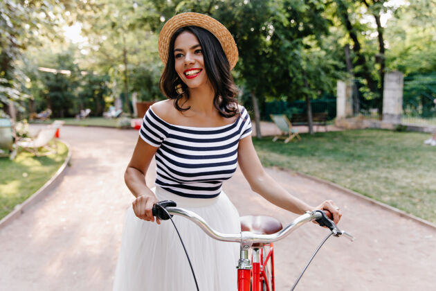 漂亮可爱的棕色头发的女孩在散步时欣赏大自然的景色美丽的拉丁女子戴着帽子在公园里和自行车合影的户外镜头微笑年轻城市