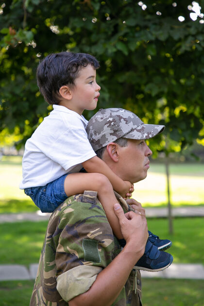 户外父亲抱着儿子在城市公园散步的侧视图高加索人的儿子穿着军装坐在父亲的脖子上 拥抱着他 向前看做父亲和回家的概念士兵前进军队