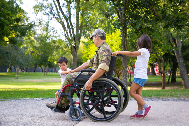 父母两个小孩和残疾军人爸爸坐在轮椅上在城市公园散步侧视图退伍军人或残疾概念军队侧视图儿子